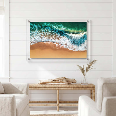 Coastal acrylic wall art with emerald water, foamy waves, and sunlit sand displayed in a bright room