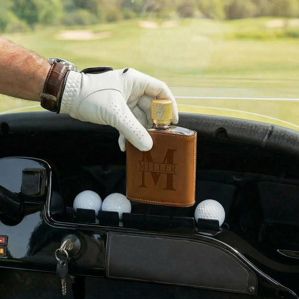 Gloved hand placing the brown leather-wrapped flask into a golf cart cup holder beside golf balls