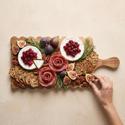 Person arranging sliced figs on a loaded scalloped acacia charcuterie board