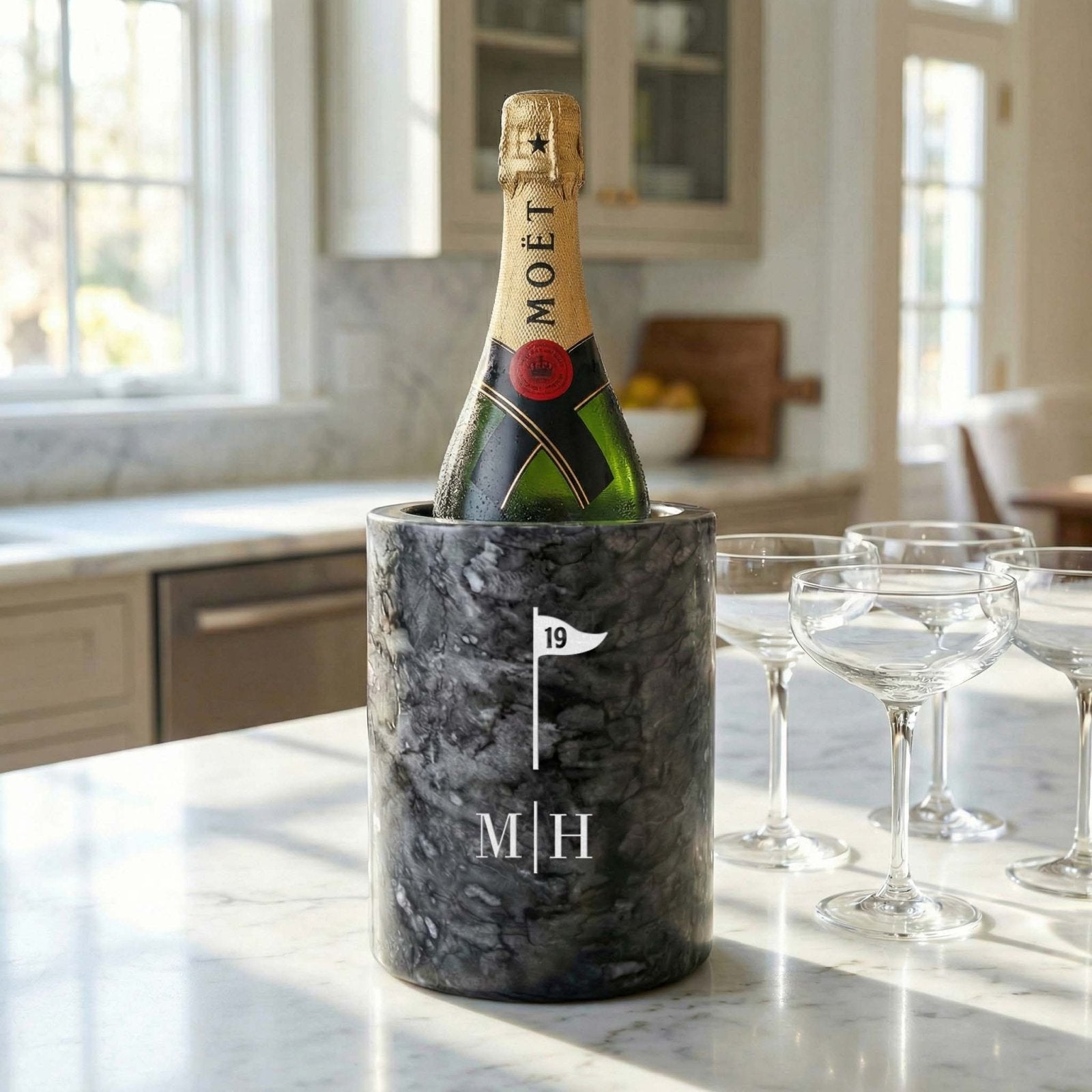 A bottle of Moët champagne in a black marble wine chiller with a golf flag icon and monogram, set on a bright marble kitchen counter beside four coupe glasses