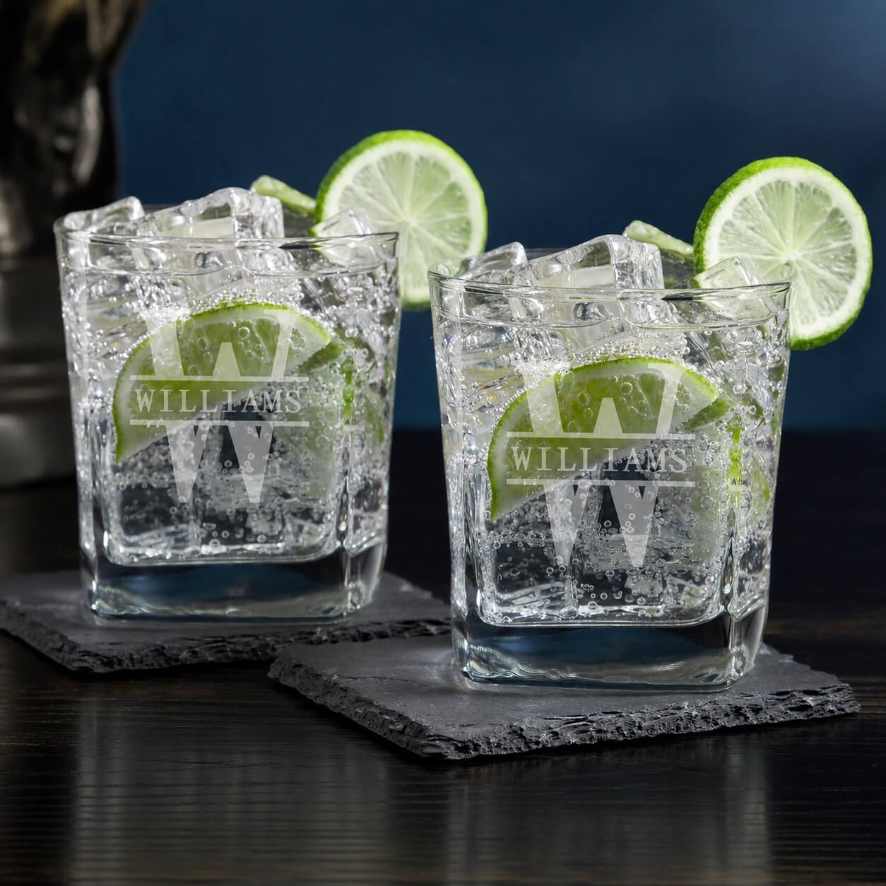 Two glasses with ice and lime wedges on dark coasters against a dark background
