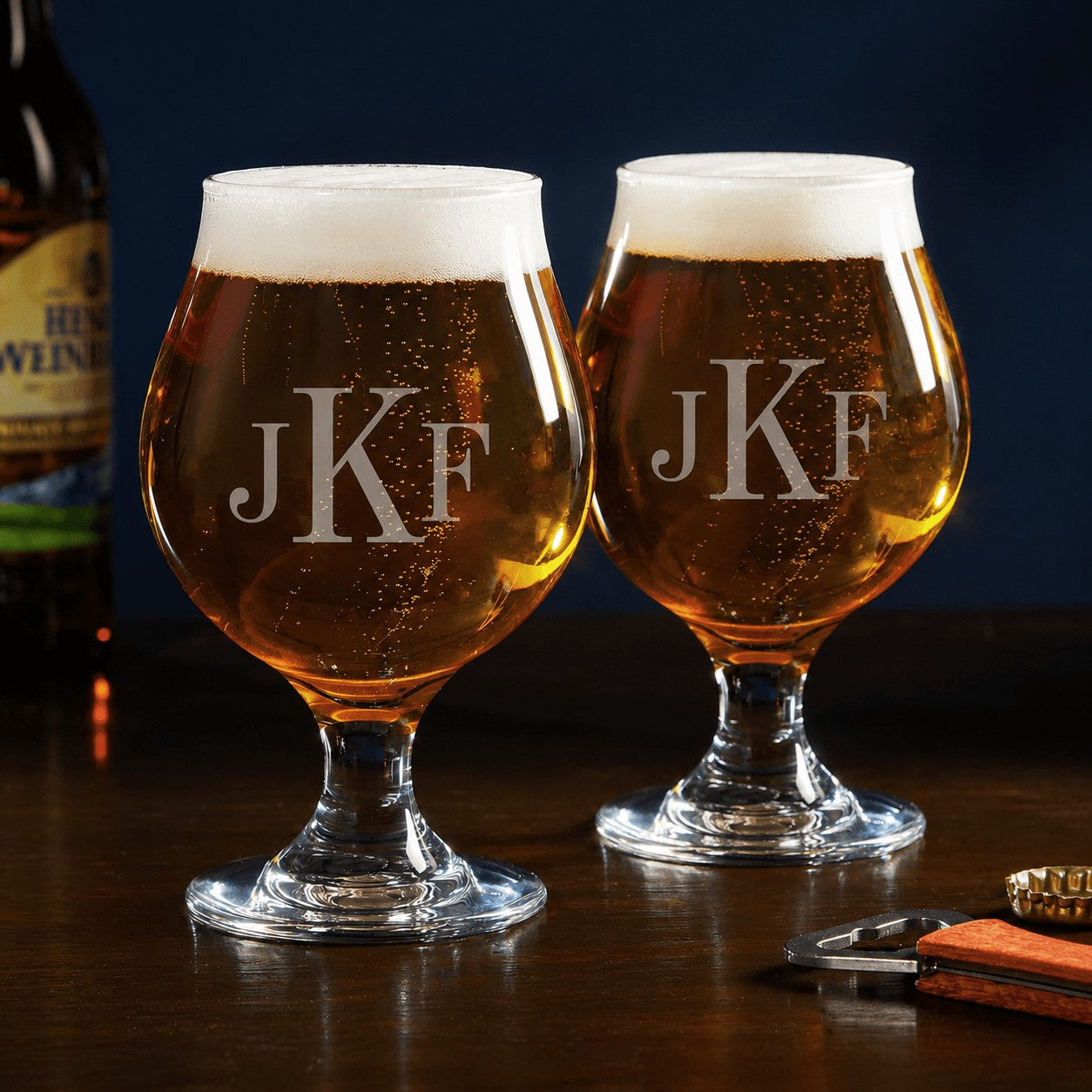Two beer glasses with 'JKF' engraving on a wooden surface with a blurred bottle in the background.