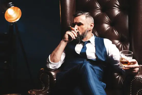 man smoking cigar holding bourbon glass on a leather couch