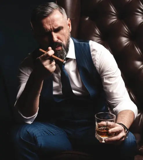 Man in suit sitting in chair drinking whiskey and smoking cigar