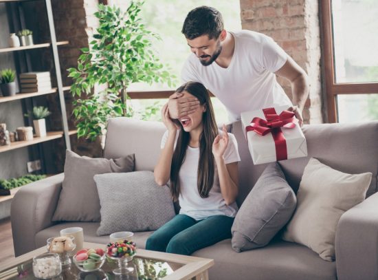 Guy surprising woman sitting on a couch with a present