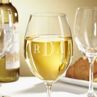Wine glass filled with white wine, monogram “RDA” on the glass; blurred bottle and other glasses in background.