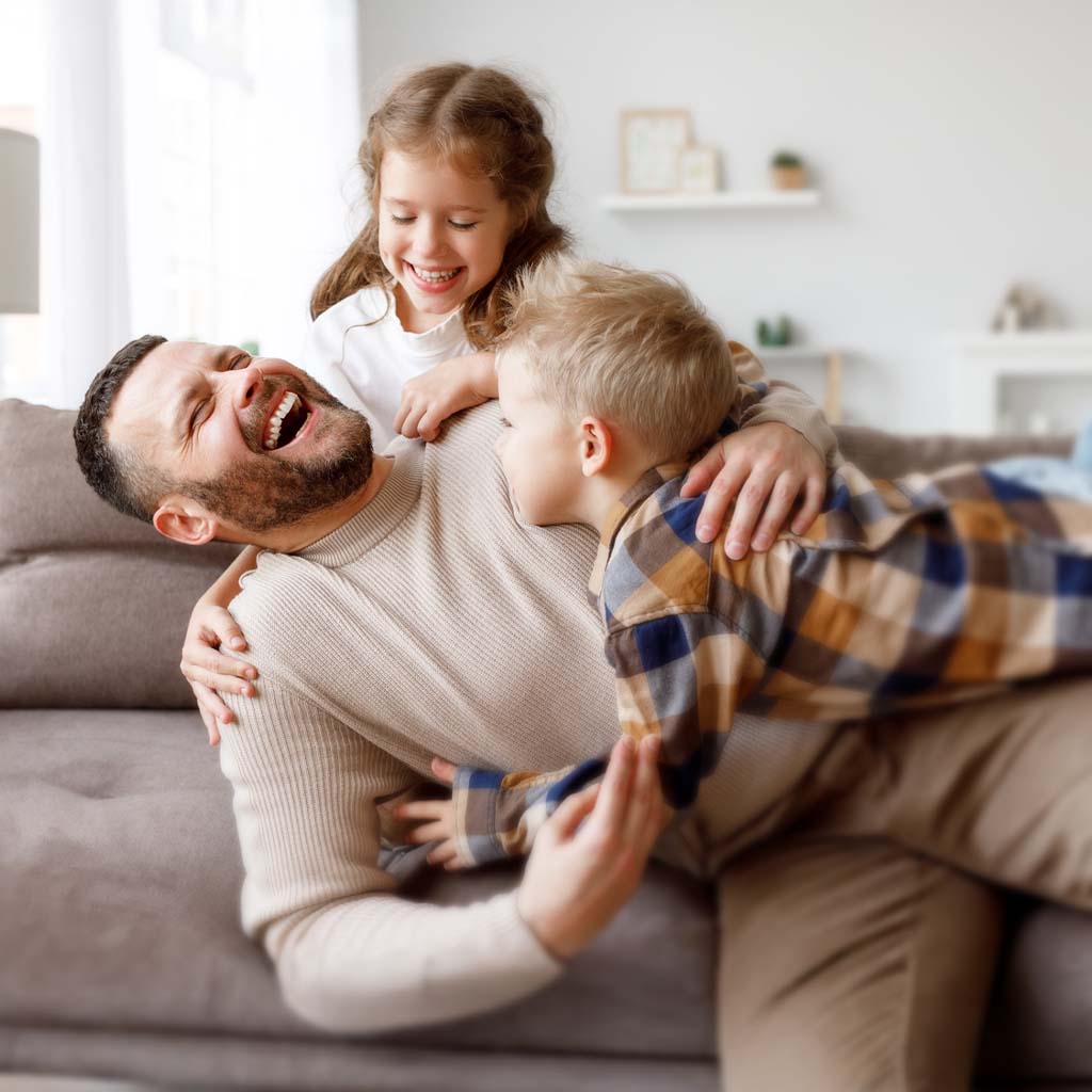 Father lying on a couch with two children, one on each side, in a bright living room.