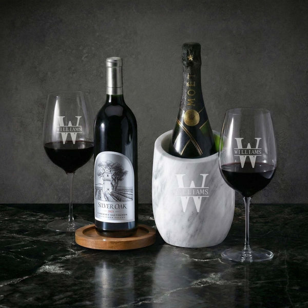 Two etched wine glasses filled with red wine sit on a dark marble surface beside a bottle of Cabernet Sauvignon. Next to it, a bottle of Moët champagne rests in a white marble wine chiller engraved with the same logo, all set against a textured gray backdrop