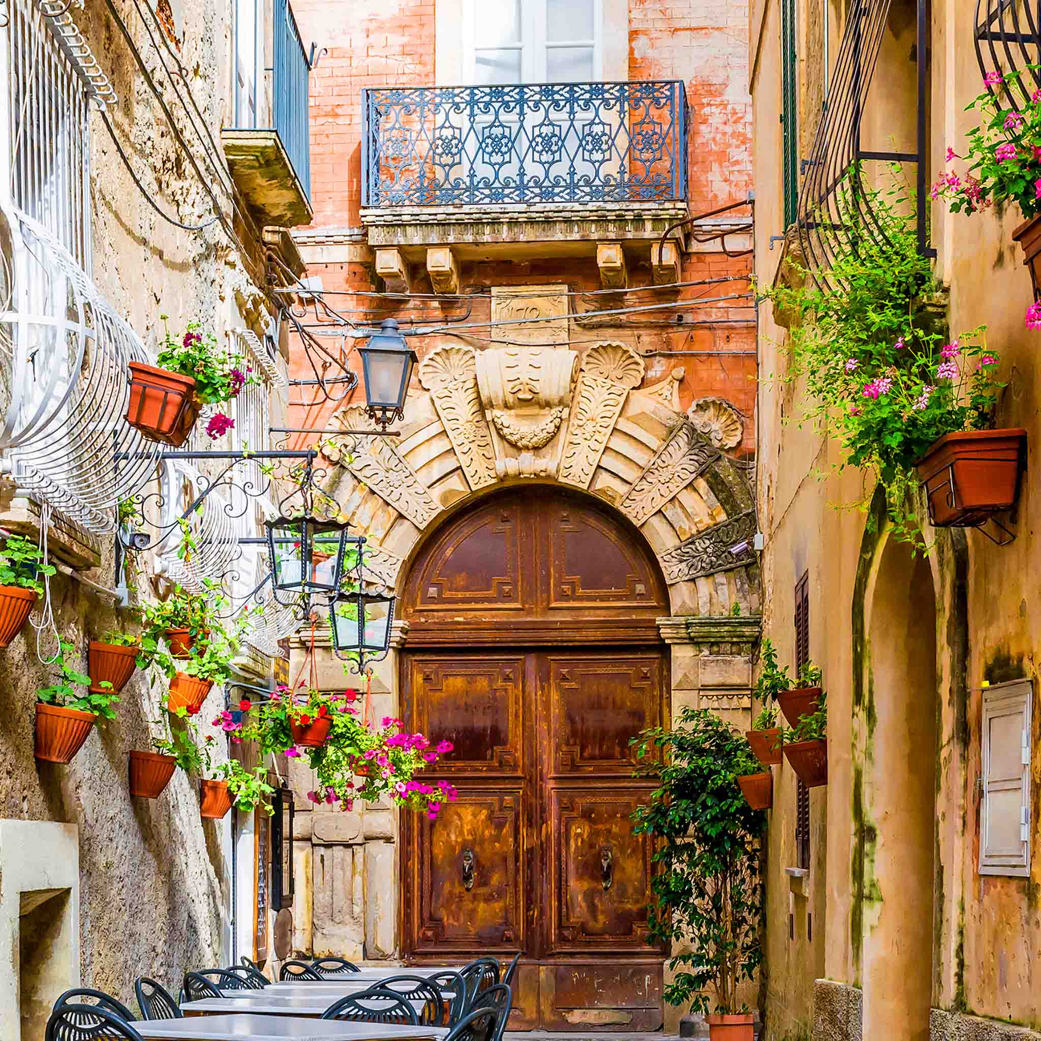 Narrow European alley lined with balconies, flowers, and outdoor seating